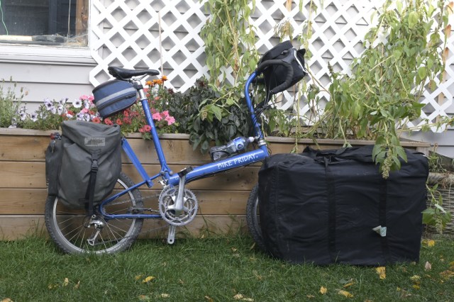 A blue folding bike parked next to a wooden planter box with flowers, accompanied by two black bags.
