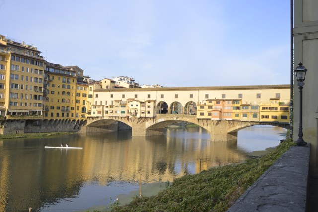 A view of the Ponte Vecchio bridge over the Arno River in Florence, Italy, with colorful buildings lining the riverbanks and a lone rower on the water.