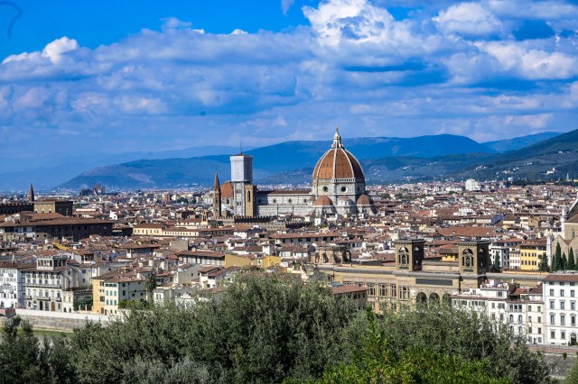 A panoramic view of Florence, Italy, featuring the iconic dome of the Duomo, surrounded by historic buildings, with hills and blue skies in the background.