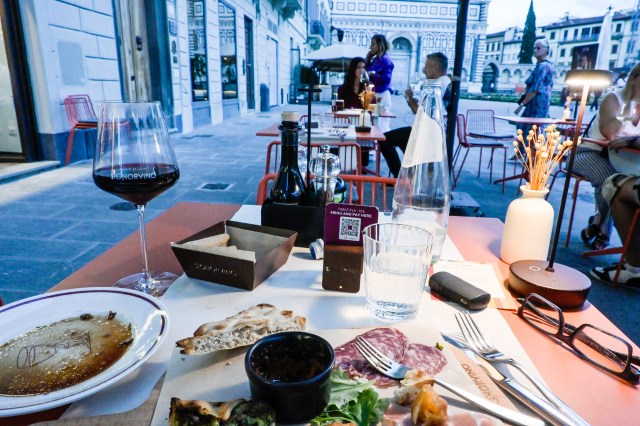 Table set for dining outdoors with a glass of red wine, a plate of food including charcuterie and bread, a bottle of water, and cutlery. Background features people seated at other tables and a view of a historic building.