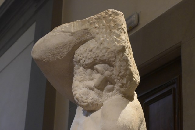Close-up of a white marble sculpture depicting a figure with a textured coat and a large wide-brimmed hat.